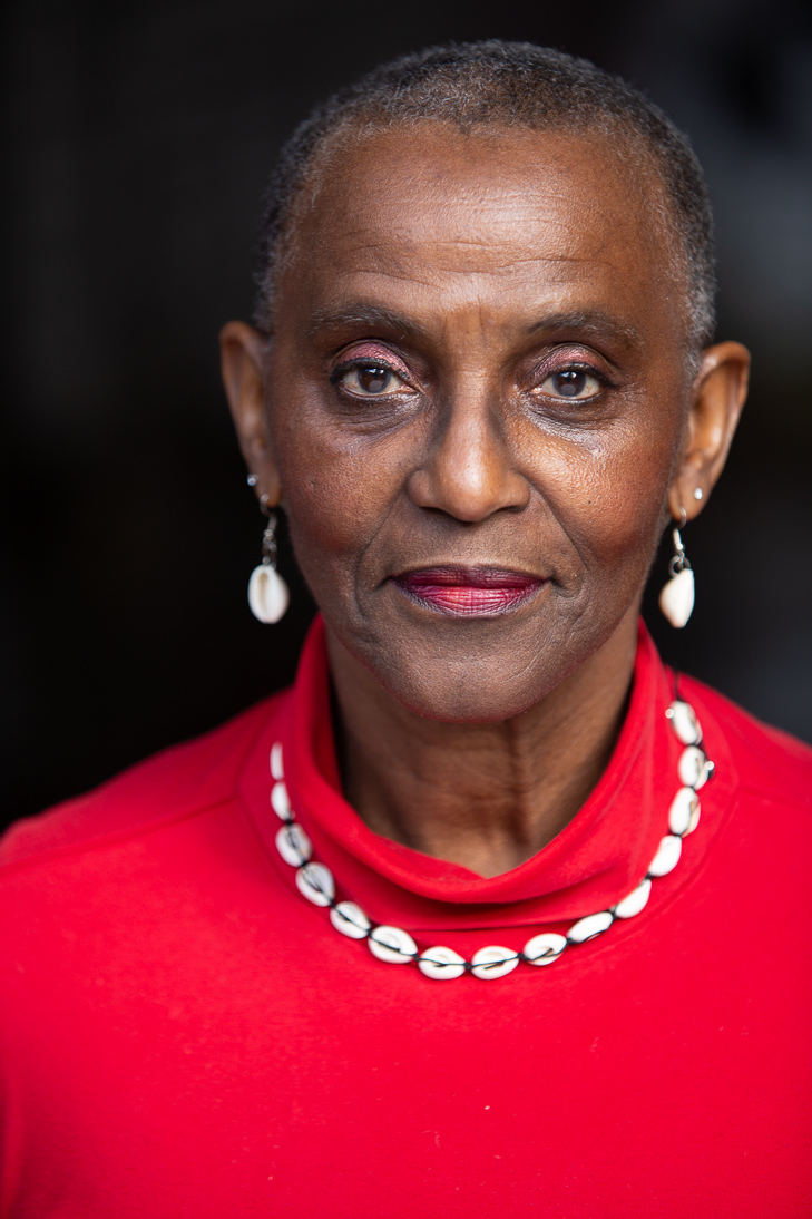 Portrait of Anni Domingo wearing a bright red top, a shell necklace, and matching shell earrings. She gazes confidently at the camera with a calm expression