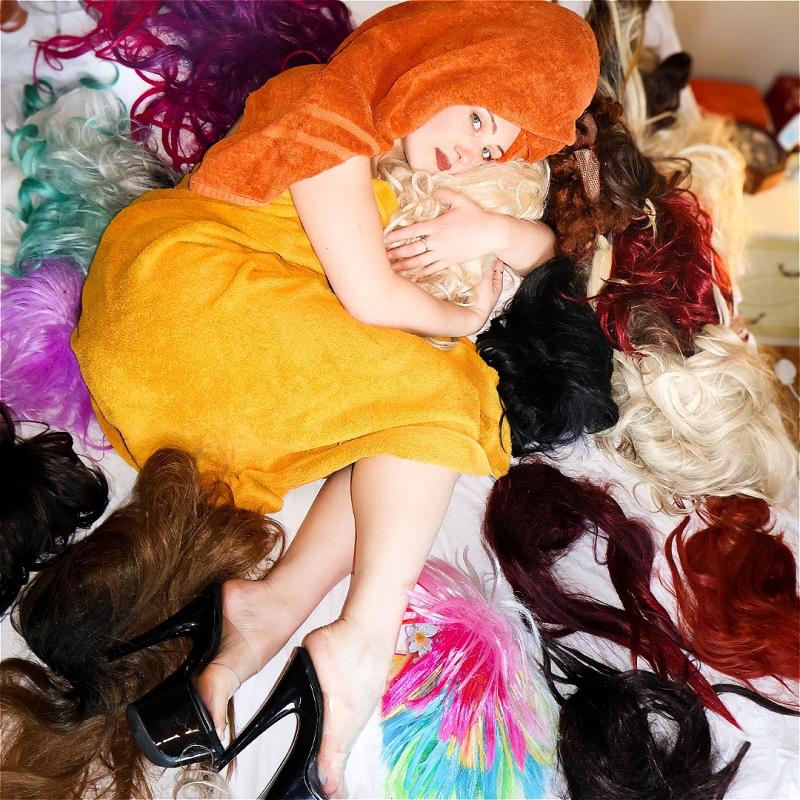 Alice Cockayne, wearing an orange towel over her head and a mustard-yellow towel wrapped around her body, lies on a bed covered in colorful wigs. She hugs a platinum blonde wig while looking at the camera, with black patent high heels on her feet