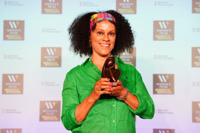 Bernardine Evaristo smiles while holding the Women’s Prize for Fiction’s bronze statuette, having received the Outstanding Contribution to Writing award. She wears a bright green shirt and a colorful patterned headband, standing in front of a backdrop featuring the Women's Prize Trust and Bukhman Philanthropies logos