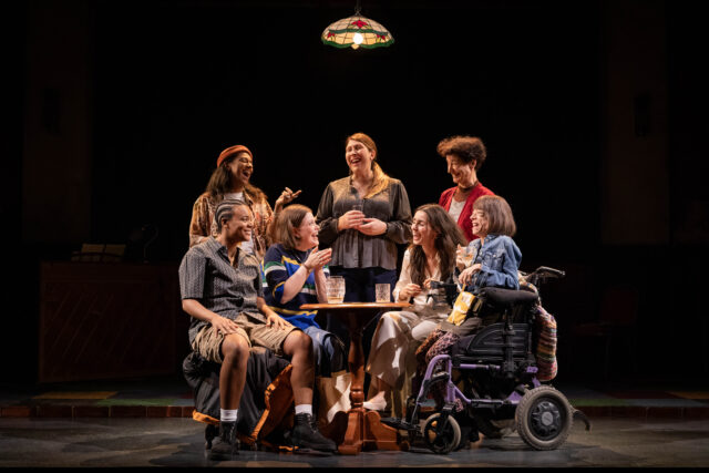 A group of seven people sit and stand around a small round table under a hanging stained glass lamp on stage in The Ministry of Lesbian Affairs at Kiln Theatre. They're laughing and mid-conversation, sharing drinks and snacks, with one woman in a wheelchair holding a cat plush toy. The warm lighting and their joyful expressions suggest a scene of camaraderie and community.