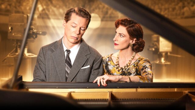 Rosalie Craig and Sean Hayes sit at a grand piano in a warmly lit studio set for Good Night, Oscar, evoking a 1950s television broadcast. Hayes, in a gray suit, looks solemn with eyes downcast, while Craig, dressed in an elegant floral dress and pearl jewelry, gazes at him with a mix of concern and intensity.