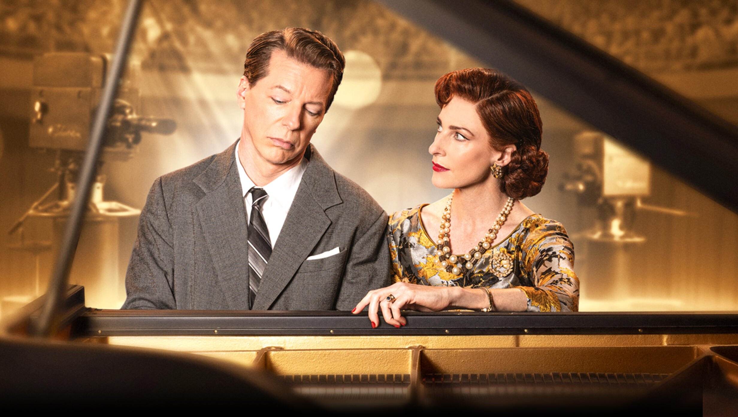 Rosalie Craig and Sean Hayes sit at a grand piano in a warmly lit studio set for Good Night, Oscar, evoking a 1950s television broadcast. Hayes, in a gray suit, looks solemn with eyes downcast, while Craig, dressed in an elegant floral dress and pearl jewelry, gazes at him with a mix of concern and intensity.