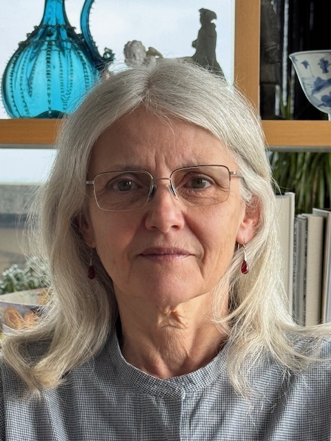 Helen Fairfoul, a woman with long gray hair and glasses, is pictured indoors looking directly at the camera with a calm expression. She wears a gray checked blouse and red drop earrings, seated in front of a wooden bookshelf decorated with books, a blue glass vase, and porcelain items. Natural light softly highlights her face.