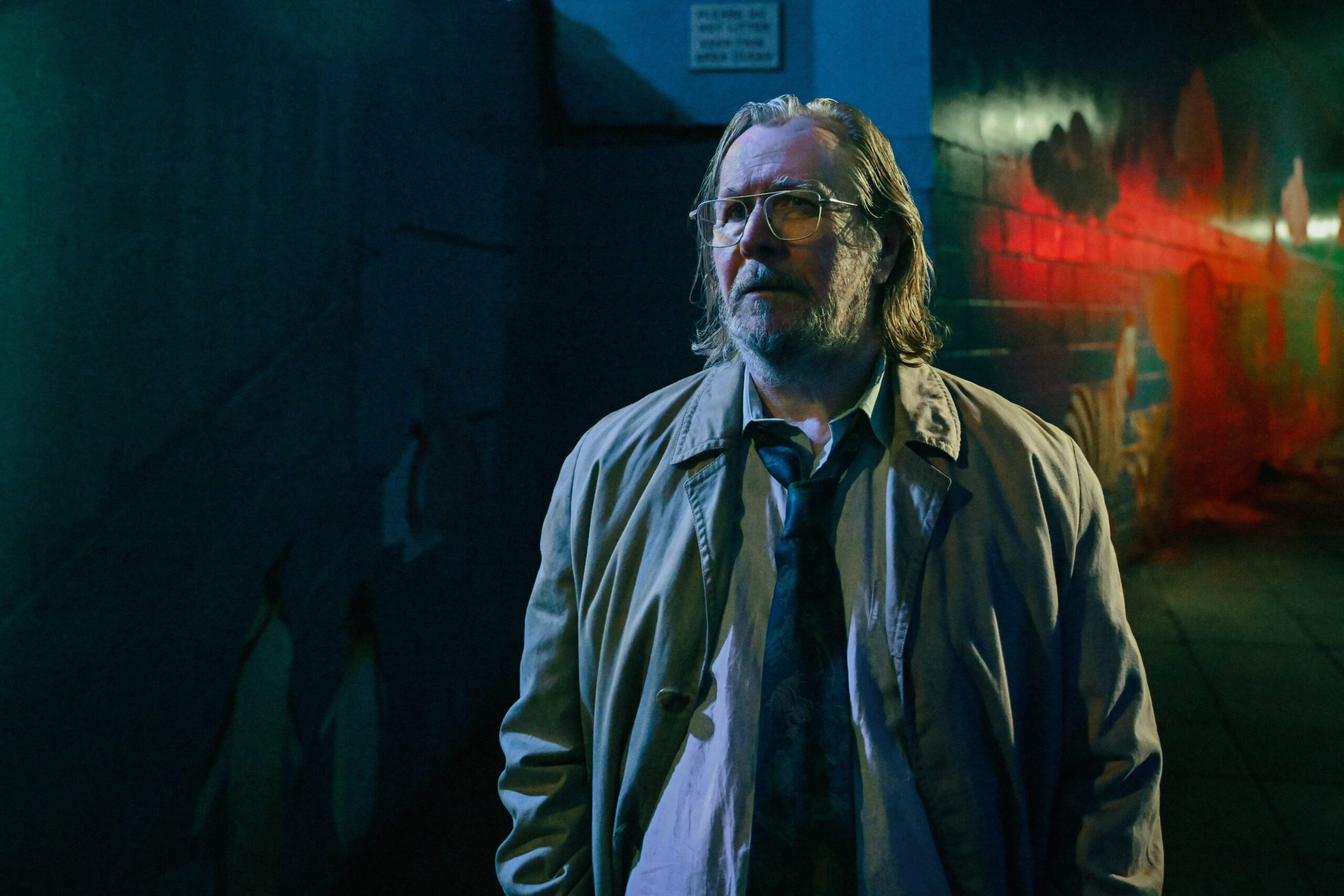 Gary Oldman stands under moody, neon-lit lighting in a trench coat and untied tie, portraying a weary expression in a gritty urban setting. This dramatic still highlights Oldman's performance in a role that has earned multiple Emmy Nominations.