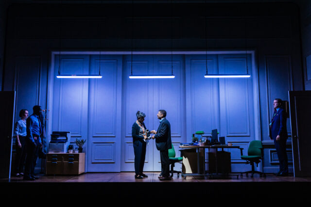 A dimly lit office stage set from The Estate at The National Theatre, bathed in blue lighting, features two actors at center stage exchanging a tense or intimate gesture under hanging fluorescent lights. On either side, office furniture, green desk chairs, and additional characters frozen in position enhance the dramatic, corporate atmosphere.