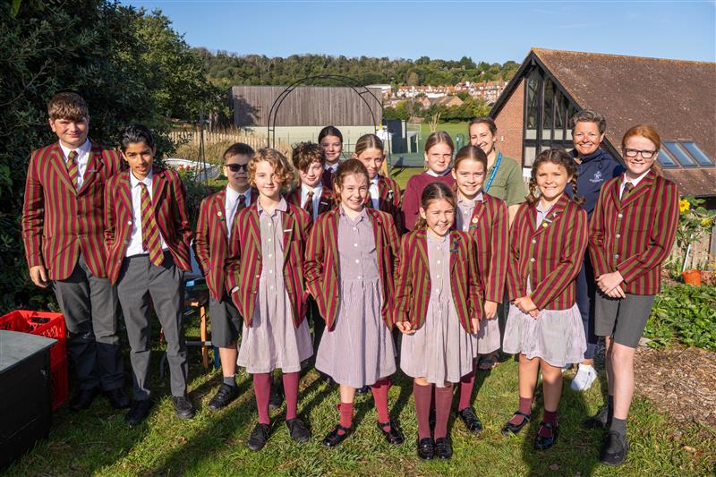 A group of St Andrew’s Prep school children in striped blazers and uniforms stand outdoors in a garden with two adults, smiling, with buildings and greenery in the background on a sunny day.