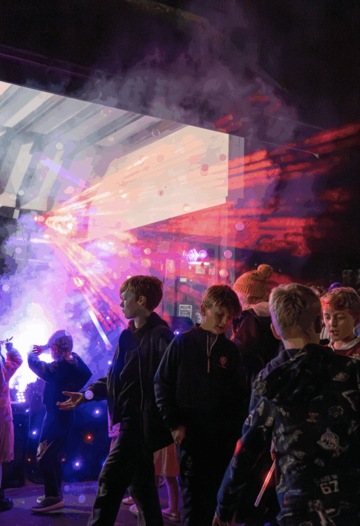A group of children stand outside at night under colourful, smoky lights. Red and purple beams and mist, like the glow of fireworks, create a festive, energetic atmosphere as the kids interact and look around.
