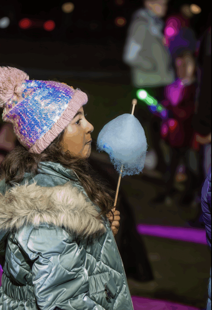 A young girl wearing a colourful beanie and a fur-trimmed coat holds blue candyfloss at an outdoor fireworks evening, with blurred people and bright lights in the background.