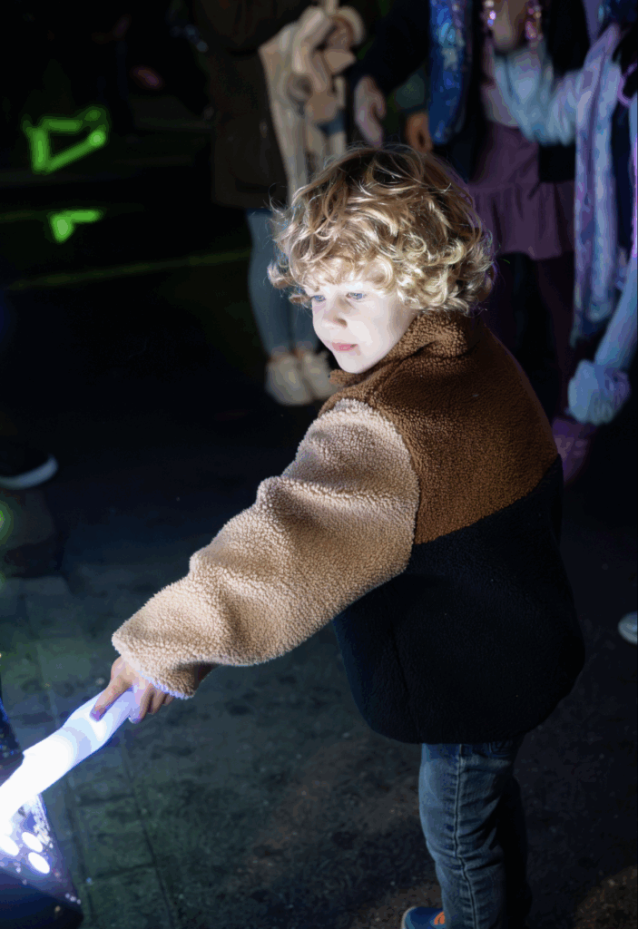 A young child with curly blond hair and a brown and black fleece jacket holds a glowing light stick on Bonfire Night, illuminated by colourful lights, with people standing in the background.