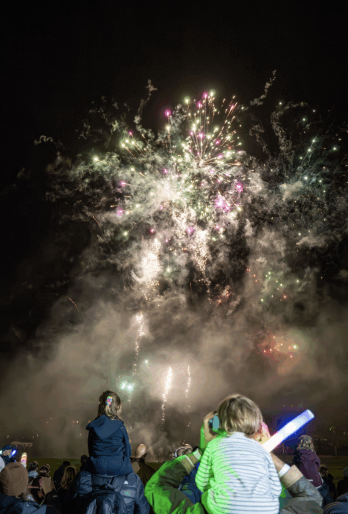 Children sit on someone's shoulders and watch colourful fireworks explode in the night sky, surrounded by mist and smoke. The kids hold glowing light sticks, making this fireworks evening a night to remember.
