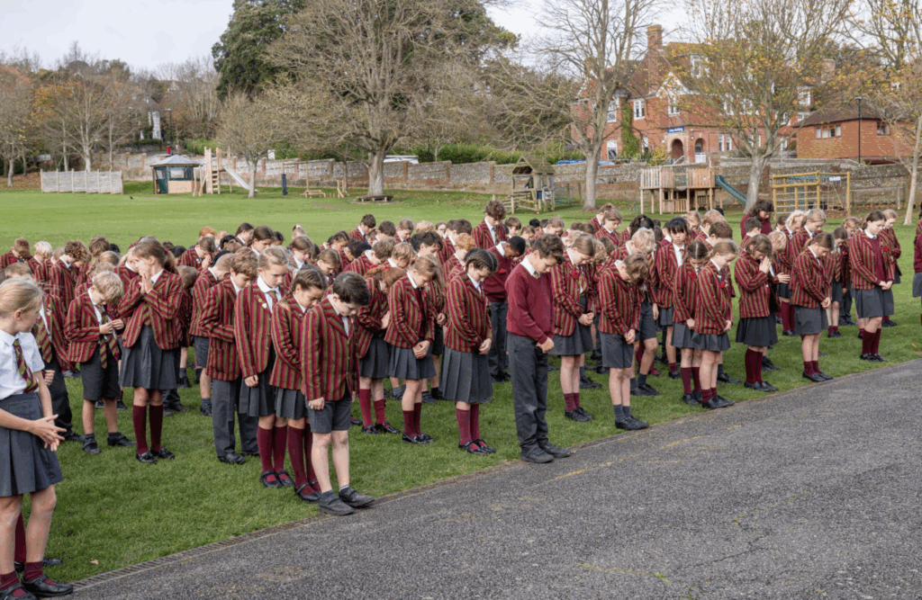 A large group of schoolchildren in striped blazers and uniforms stand outdoors on grass with heads bowed, likely participating in a Time for Reflection. Trees and brick buildings can be seen in the background.