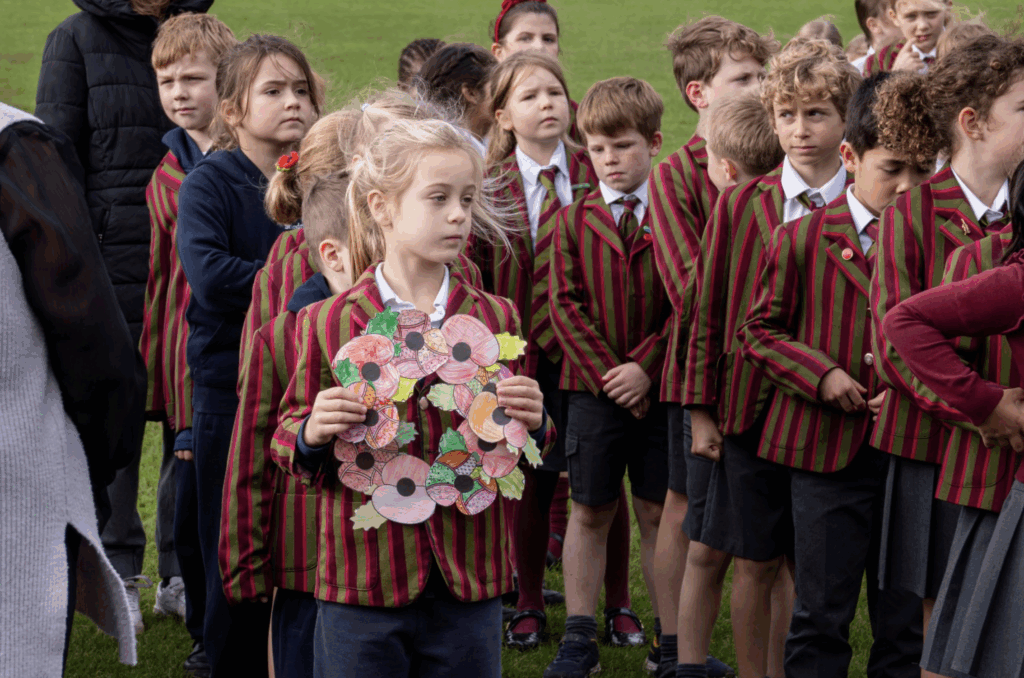 A group of schoolchildren in red and green striped blazers and ties stand outdoors. A girl at the front holds a handmade paper wreath decorated with red poppies, as the children gather for a ceremony—a time for reflection and remembrance.