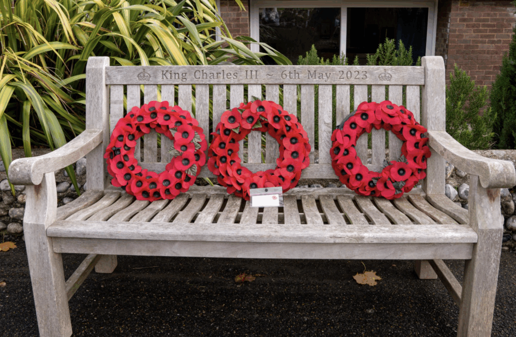 A wooden bench engraved with King Charles III 6th May 2023 holds three red poppy wreaths and a small white card, offering a time for reflection outdoors near green plants and a brick building.