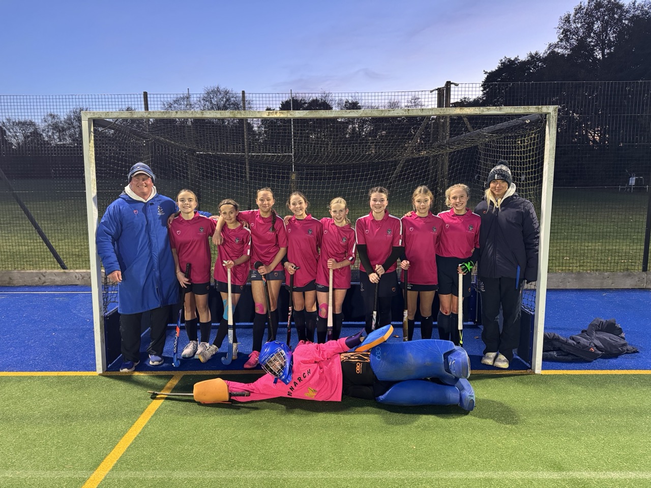 A girls’ hockey team in pink jerseys, the Girls 1st XI, poses with two coaches and their blue-clad goalkeeper at the IAPS National Finals, where they earned Overall 4th. It’s evening on an outdoor artificial pitch with trees in the background.
