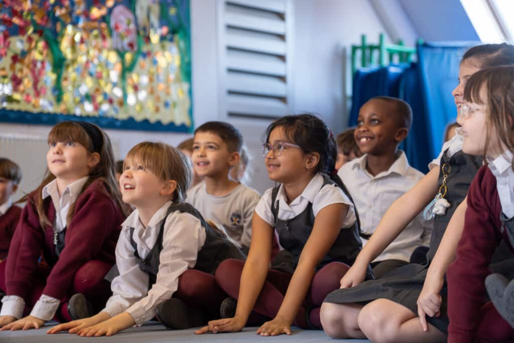 A group of young schoolchildren in uniforms sit on the floor indoors at St Andrew’s Prep, smiling and looking ahead with interest. The background displays colourful artwork and classroom elements typical of top UK prep schools.