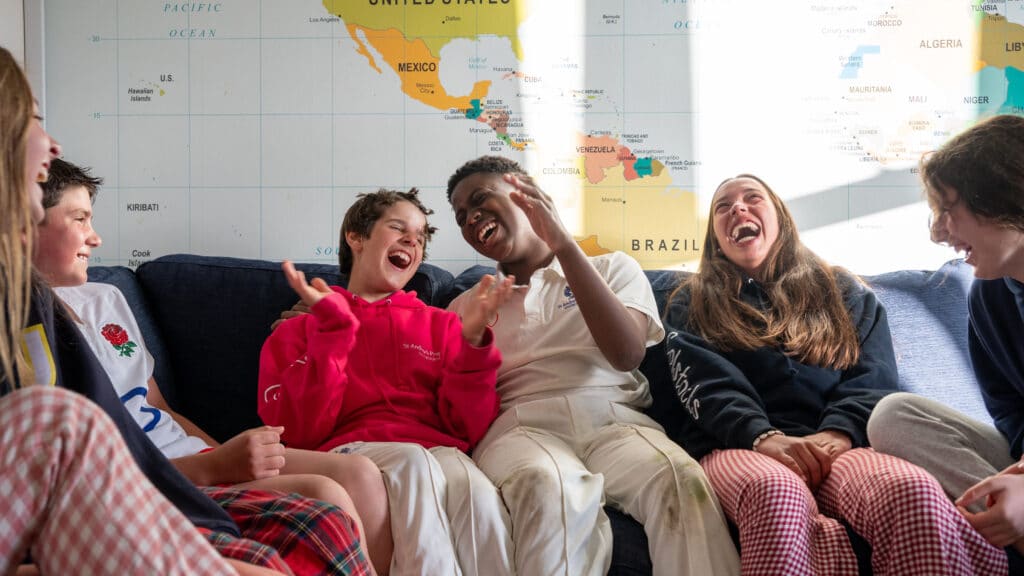 A group of six teenagers from St Andrew's Prep sit on a sofa, laughing and talking together. Behind them, a large world map highlights the Americas whilst sunlight brightens the welcoming prep school boarding scene.
