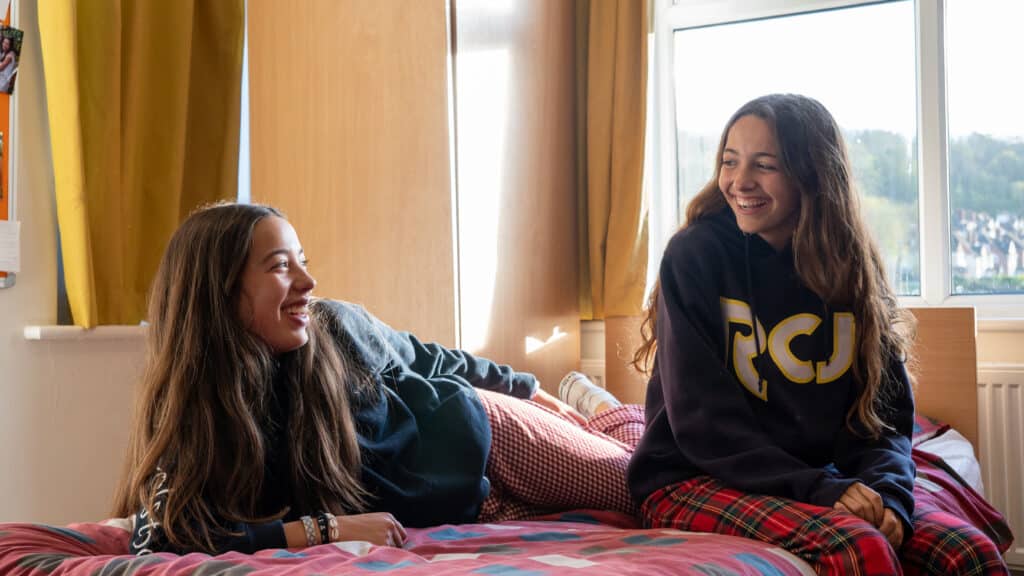 Two young women with long hair, wearing casual clothes, sit on a bed and smile as they chat about St Andrew’s Prep in a brightly lit room with yellow curtains and a window, reflecting the friendly atmosphere of prep school boarding.