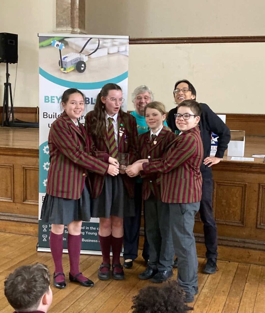 Four St Andrew’s Pupils in maroon-striped uniforms smile proudly with a trophy after the Young Engineers Challenge 2026, as three adults stand behind them and a banner about building robots is visible in the background.