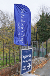 A blue flag reading “St Andrew’s Prep Eastbourne” stands beside a sign pointing right that says “St Andrew’s Prep Netball Festival and U11 Football this way.” Trees with autumn leaves are in the background.