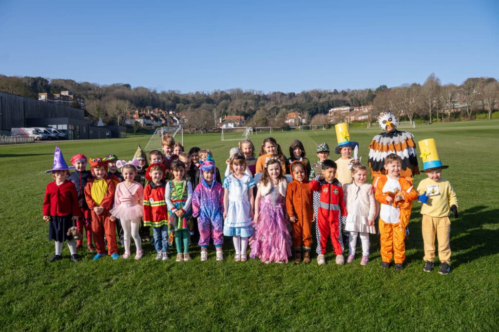 A group of young children in colourful costumes stand in a row on a grassy field with trees and houses in the background, smiling and posing for a cheerful World Book Day 2026 photo on a sunny day.