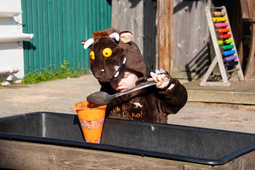 A young child dressed in a Gruffalo costume celebrates World Book Day 2026 by using a large spoon to scoop mud into an orange funnel at an outdoor play area. A colourful xylophone stands in the background.