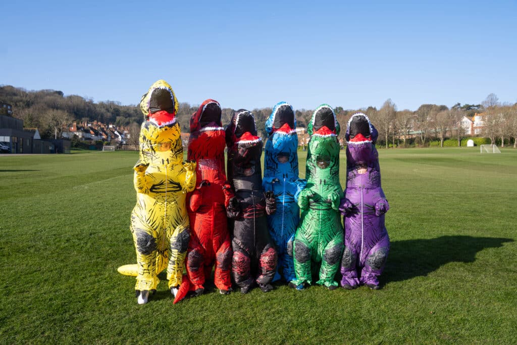 Seven people wearing inflatable dinosaur costumes in different colours—yellow, orange, red, black, blue, green, and purple—stand together on a grassy field under a clear blue sky celebrating World Book Day 2026.