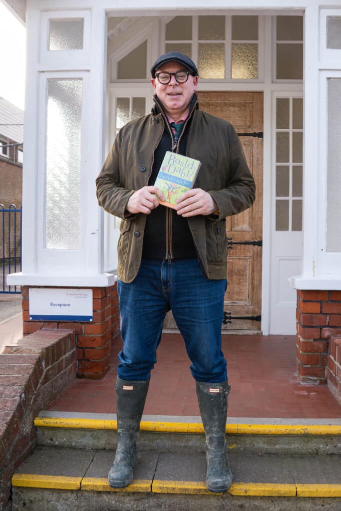 A man wearing glasses, a flat cap, a brown jacket, jeans, and muddy boots stands on a doorstep holding a Roald Dahl book and smiling at the camera for World Book Day 2026.