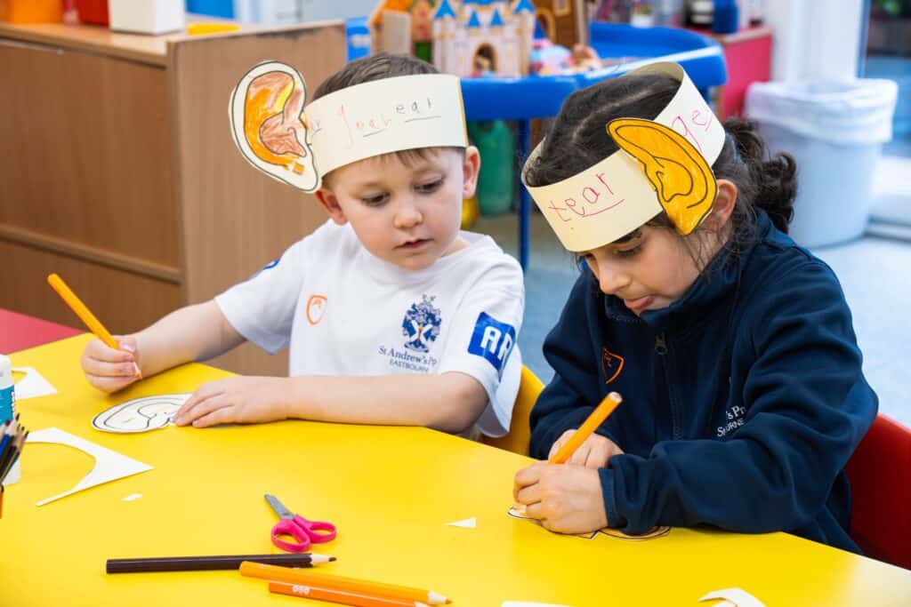 Two young children sit at a yellow table in a classroom, colouring ear shapes for their study. They wear headbands labelled "ear," each with a paper ear attached. Colourful pencils, scissors, and paper are scattered on the table as the Headteacher observes nearby.