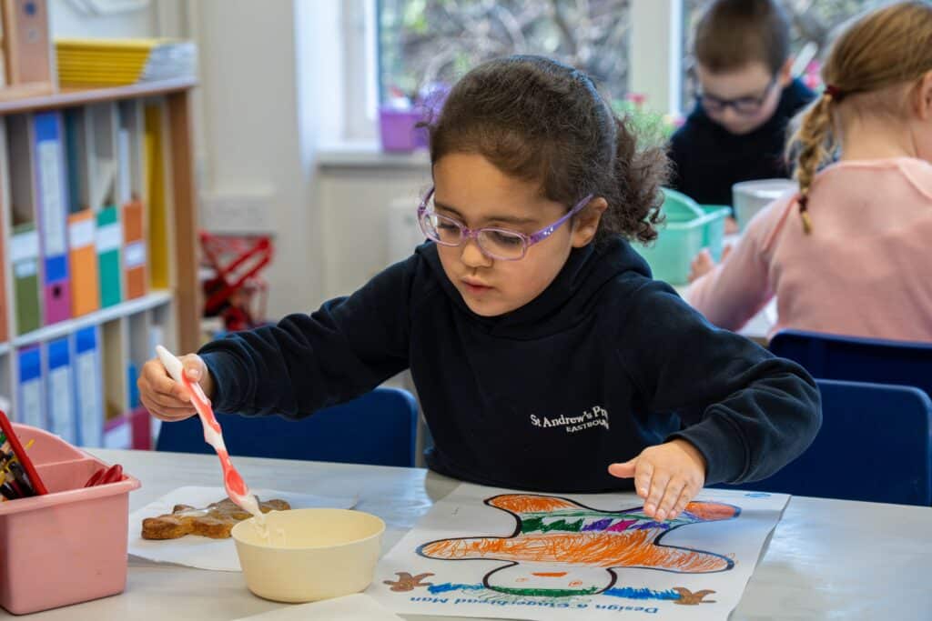 A young girl with glasses sits at a table, using a paintbrush to decorate a gingerbread man colouring sheet as part of her study. Other children are also working in the background. The classroom is bright and organised, overseen by the headteacher.