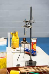 A chemistry experiment set-up for Science Week features a test tube clamped above a small burner, surrounded by bags of food items on a white table in a classroom or laboratory setting.