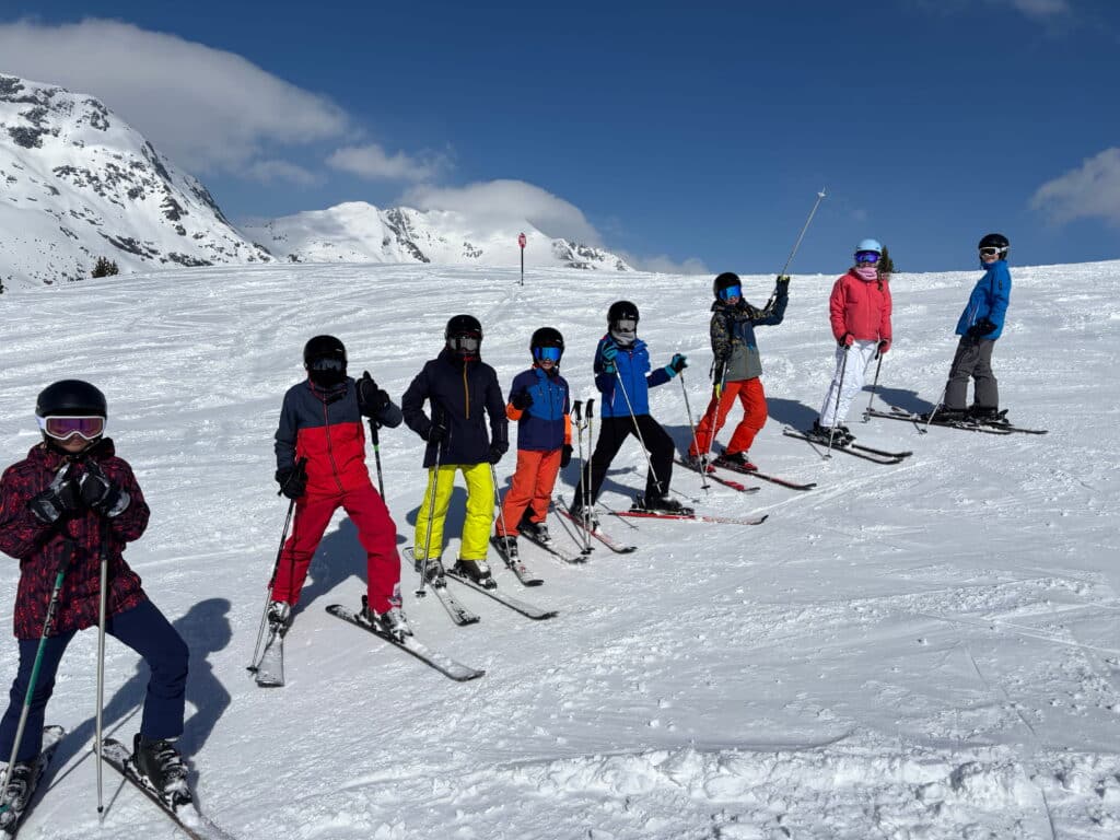 A group of seven children in colourful ski gear stand in a line on the Andorran slopes, holding ski poles, with snow-covered peaks and a blue sky in the background, showcasing teamwork on the mountain.