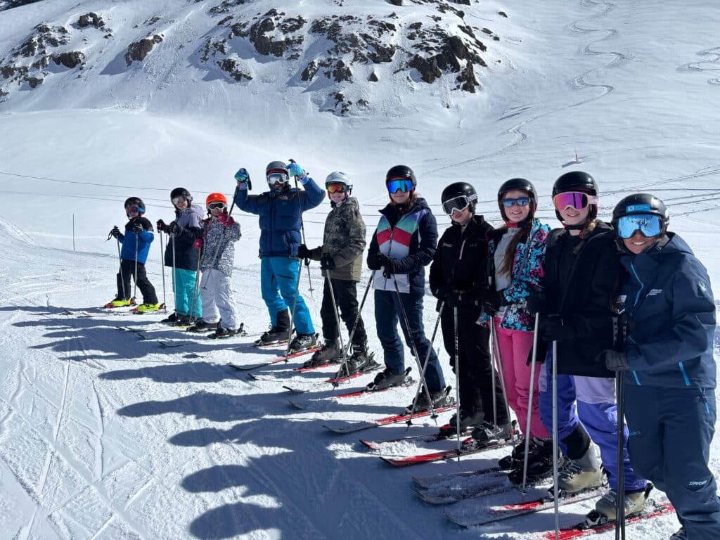A group of skiers wearing helmets, goggles, and winter gear stand in a queue on Andorran slopes, posing for a photo under bright sunlight with snow-covered rocks in the background, showcasing teamwork and adventure.
