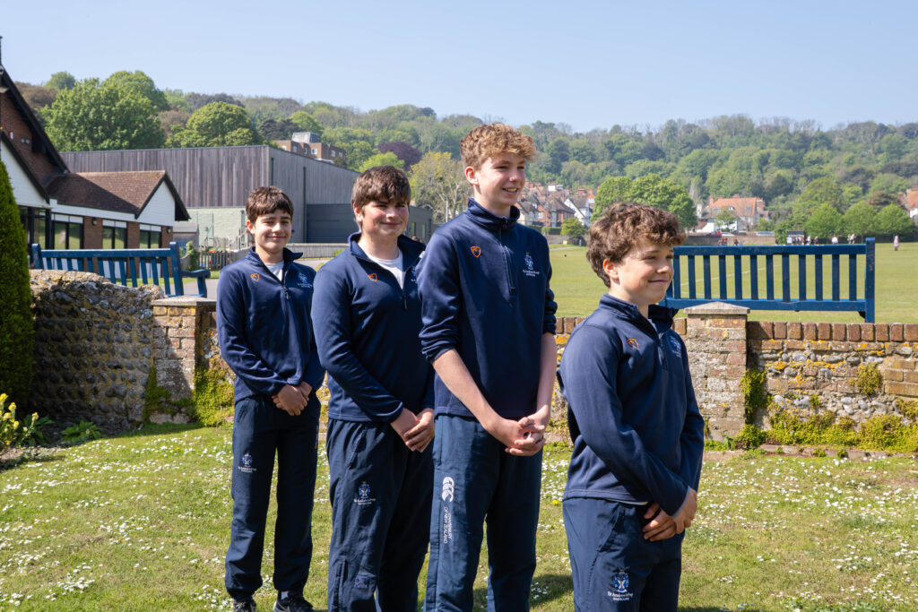 Four boys in matching navy sports uniforms from St Andrew’s Prep stand in a queue on a grassy area, with buildings, a fence, and green trees behind them on a bright, sunny day at the 2026 IAPS National Finals.