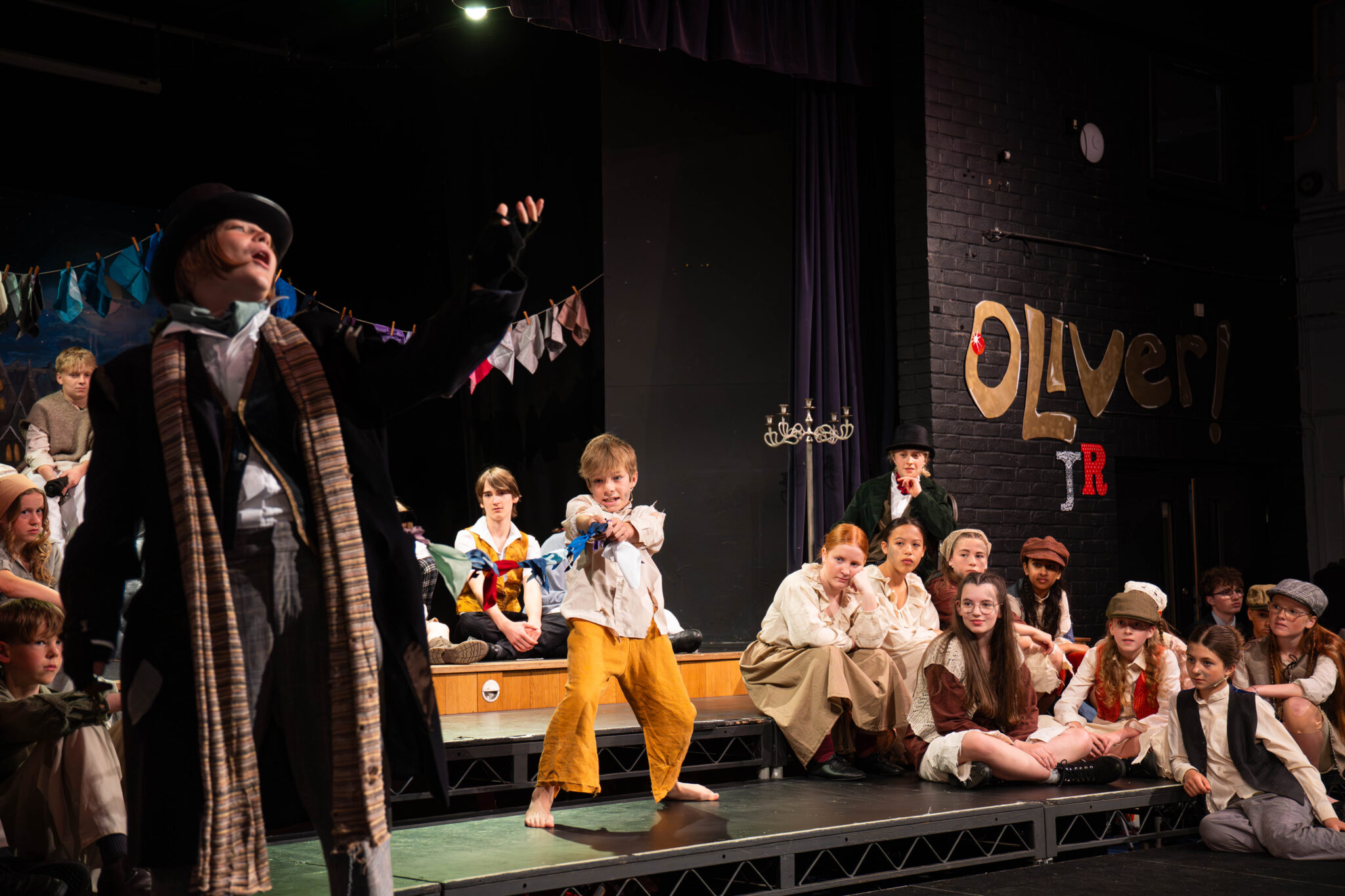 Children perform in a stage production of Oliver! One child in yellow trousers stands centre stage, gesturing, while others in period costumes sit and watch. A handmade Oliver! sign hangs on the black brick wall.